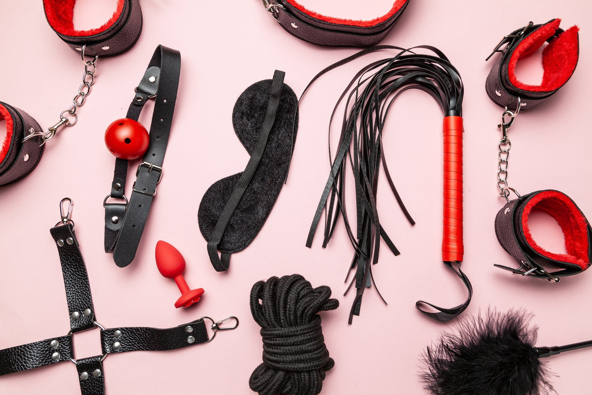 BDSM accessories including restraints, a blindfold, rope, flogger, ball gag, and plug arranged on a pink background. Featured in a guide explaining what BDSM is and common beginner gear.