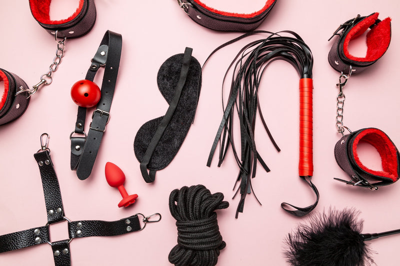 BDSM accessories including restraints, a blindfold, rope, flogger, ball gag, and plug arranged on a pink background. Featured in a guide explaining what BDSM is and common beginner gear.