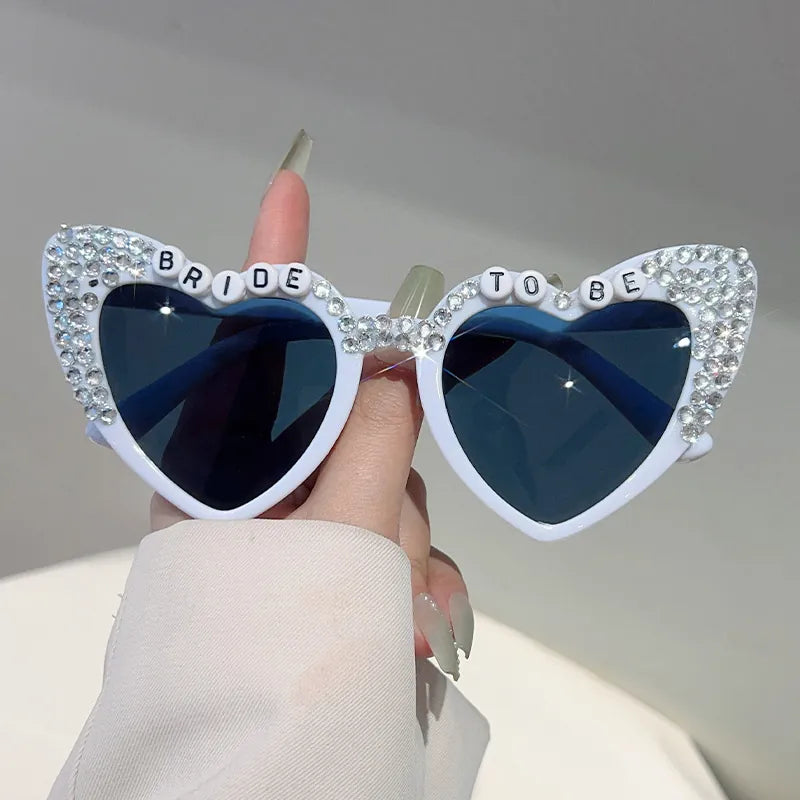 White heart-shaped sunglasses with rhinestone Bride To Be lettering and grey lenses